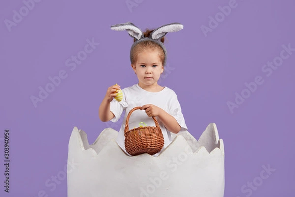 Fototapeta Happy child girl in costume Easter bunny rabbit with ears and a basket with eggs. Little girl in a big chicken egg on a purple background