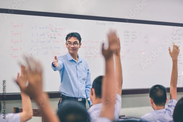 Fototapeta An smiling Asian male high school teacher teaches the white uniform students in the classroom by asking questions and then the students raise their hands for answers.