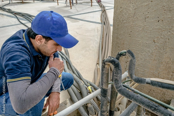 Fototapeta a professional electrician man is fixing the heavy unit of an air conditioner at the roof top of a building and wearing blue uniform and head cap