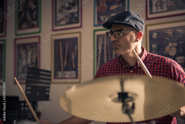 Fototapeta Caucasian young man with glasses and beret wearing a red and black plaid shirt, playing the drums with a soft side light
