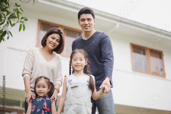Fototapeta Young Asian family love, father mother and two daughter standing at outdoor in front of home. girl and parent looking at camera which smiling and felling happy and background is a white house.