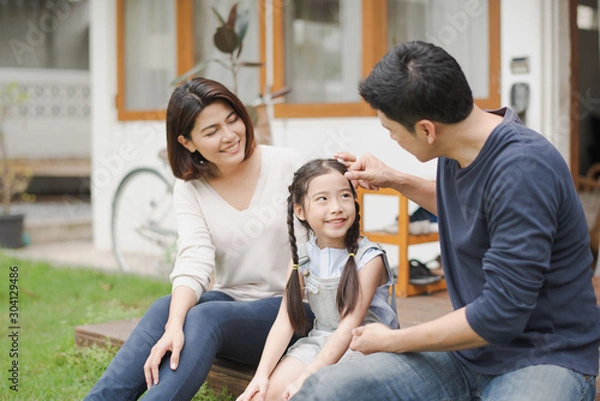 Fototapeta Young Asian family love, father mother and daughter sitting talking smiling at grass outdoor in front of home. girl looking at father which smiling and felling happy. background is white house.