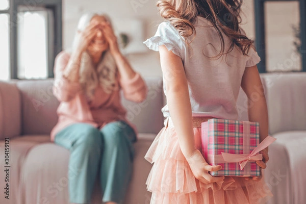 Fototapeta girl holding present box in the hands behind her back and making surprise for grandmother sitting on the couch