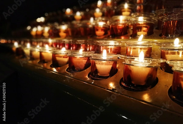 Fototapeta Candles in a church, cathedral or temple, in yellow transparent candlesticks. The concept of mourning. We remember, we grieve. Selective focus, side view, copy space