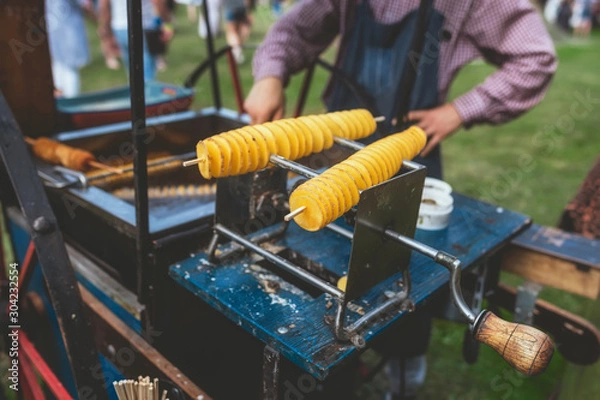 Fototapeta Spiral deep-fried potato chips on sticks in a street food vendor stall during outdoor event. A man frying and selling tornado potatoes. Street food concept. Close-up, toned