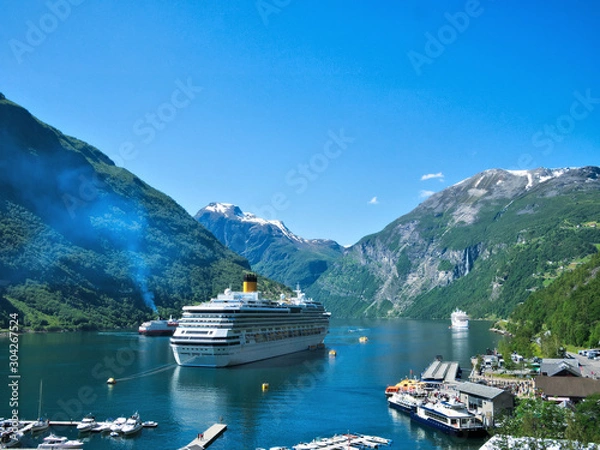 Obraz Spectacular view of Geiranger Fjord and mountains with snow, waterfalls, moored cruise ships and tender boats carrying tourists from ship to shore on a beautiful day with blue sky.