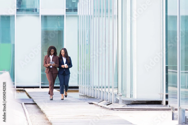 Fototapeta Full length portrait of smiling businesswomen walking with tablet. Cheerful young women wearing formal wear looking at tablet during stroll. Business concept