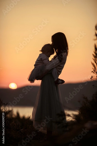Fototapeta Silhouettes of mother with a baby in her arms on nature with a view of the sun setting in the water, evening at golden hour