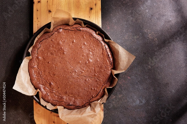 Fototapeta Freshly baked homemade chocolate cake - brownie in round form pen with paper on rustic black background. Top view, flat lay, copy space.  Light from window. 