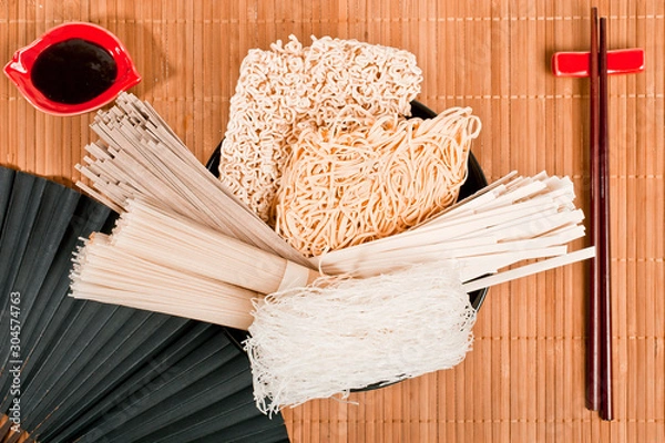 Fototapeta Different bundles of uncooked asian noodles (udon, soba, ramen, rice and glass noodles) in black clay bowl with soy sauce and black paper fan, close up on bamboo background. Top view.