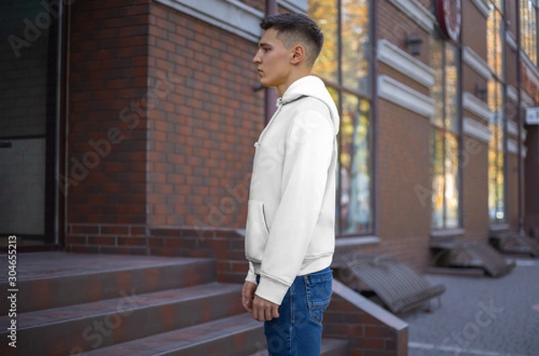 Fototapeta Side view of a white hoodie presentation on a young guy against the background of a brick building.