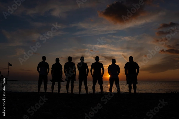 Fototapeta Silhouettes of men standing by the sea against the sunset. Group of unrecognizable men with descending sun behind their bodies.