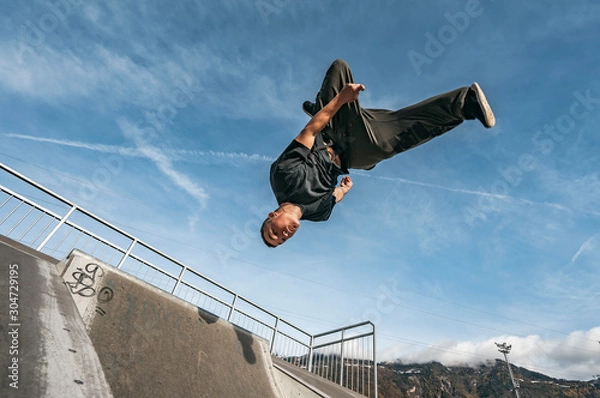 Fototapeta Young Parkour and Freerunning athlet doing a backflip from a wall in an urban enviroment with a blue sky in de background, jumping tumbling  Gymnastics training concept
