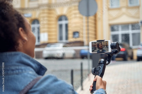 Fototapeta Outdoors Leisure. Young girl blogger walking with stabilizer recording vlog on smartphone talking to camera joyful back view close-up