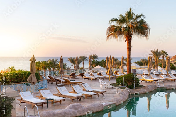 Obraz Pool early in the morning at sunrise. Sea view from an exotic resort. Nobody at the pool, sunbeds are waiting for guests. Sea horizon at sunrise.