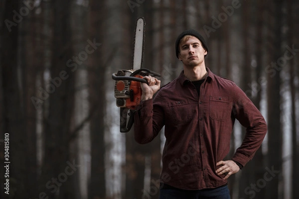 Fototapeta Lumberman working with chainsaw in the forest. Strong lumberjack with the chainsow in the forest.Stylish lumberman getting ready for work. Lifestyle.
