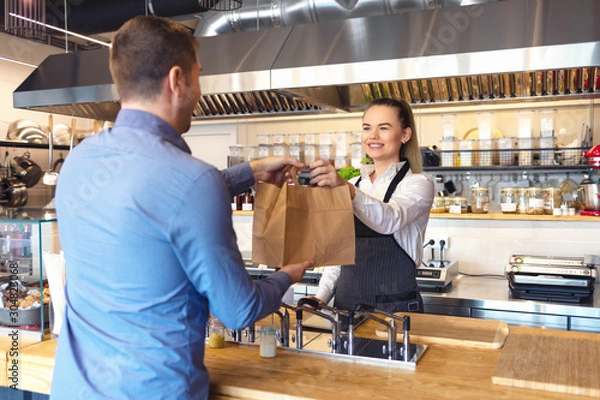 Fototapeta Happy waitress wearing apron serving takeaway food to customer at counter in small family restaurant. 