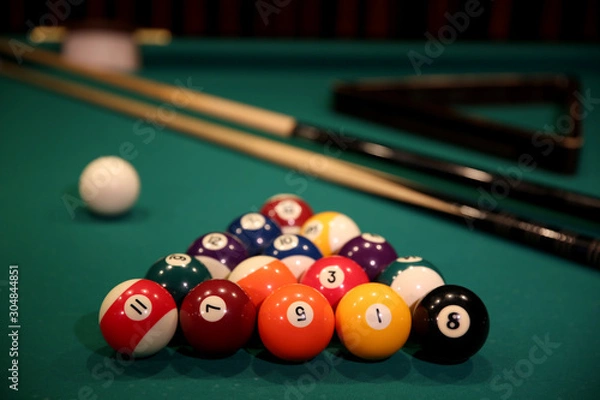 Fototapeta Sports game of billiards on a green cloth. Multi-colored billiard balls in the form of a triangle with numbers, two cues, a cue ball and a triangle on a pool table. Billiards billiard balls close up.