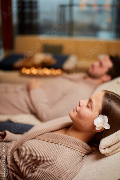 Obraz Young caucasian couple take rest in spa salon lying in bathrobes together and relax with candles