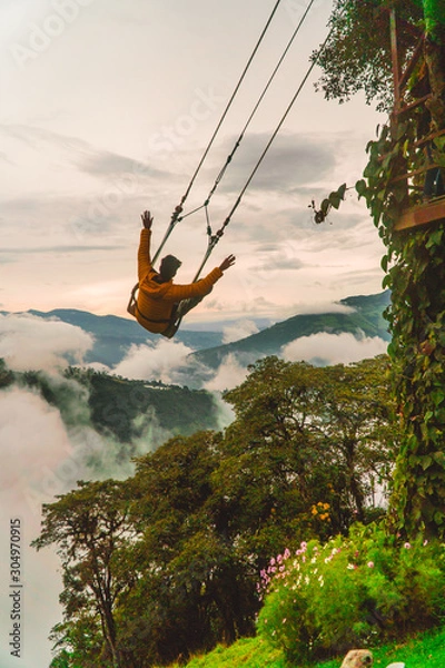 Fototapeta Man on giant swing Ecuador. Tourist having fun swinging on Giant Swing in Casa del Arbol attraction. View of green mountains and trees in background. Wooden Treehouse. Shot in Banos, Ecuador