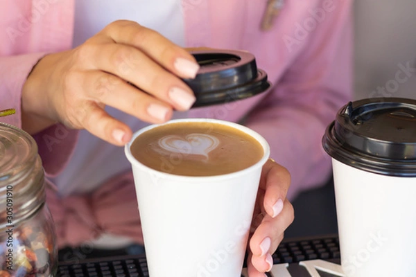 Fototapeta Barista woman closes a mug with a coffee lid. Cappuccino with a heart-shaped pattern. Closeup