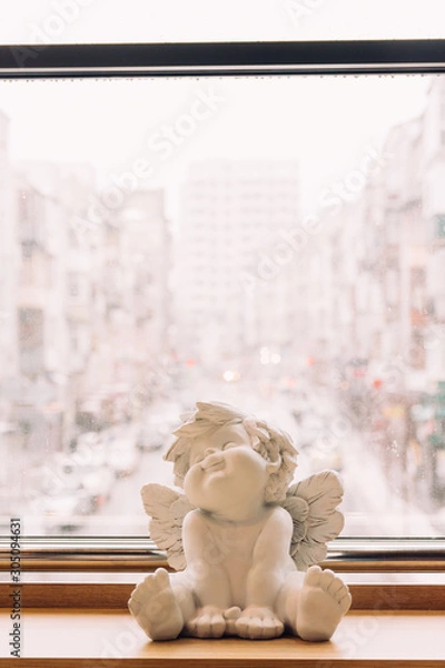 Obraz sculpture of an angel, decor, stands on the background of the city, an angel sits on a wooden table, windowsill. Valentine's Day. protection. Guardian