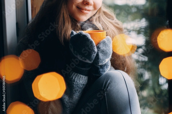 Obraz Woman's hand holding a cup of coffee or cocoain in gloves sitting home by the window. Blurred winter snow tree background. Morning, coziness, winter and people concept