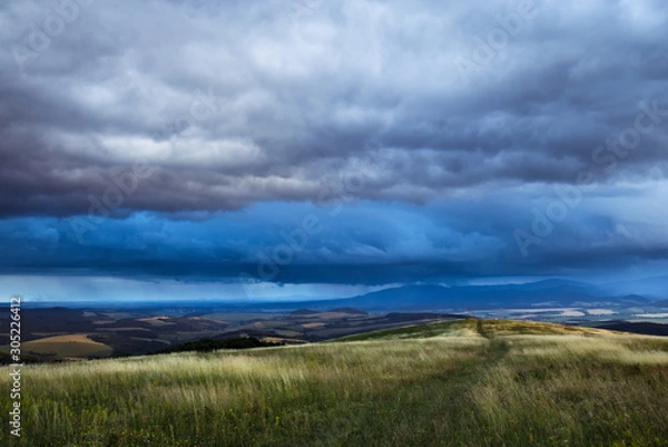 Fototapeta A grassy meadow with a road and a view under a stormy sky