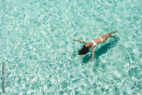 Fototapeta Woman in white bikini lying on transparent turquoise water surface on beach. Travel and vacations concept. Tropical background with empty space