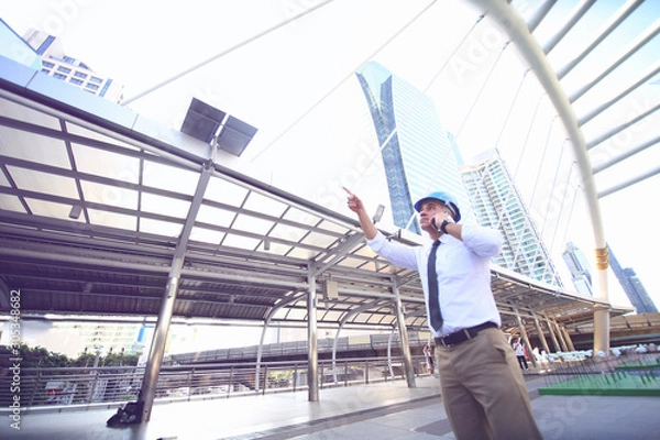 Fototapeta The engineer holding blueprint standing against  building. The concept of engineering, construction, city life and future.