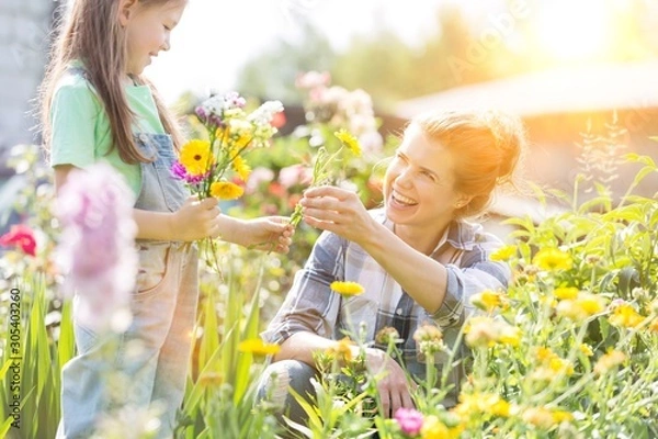 Fototapeta Smiling mother giving flowers to daughter while gardening at farm