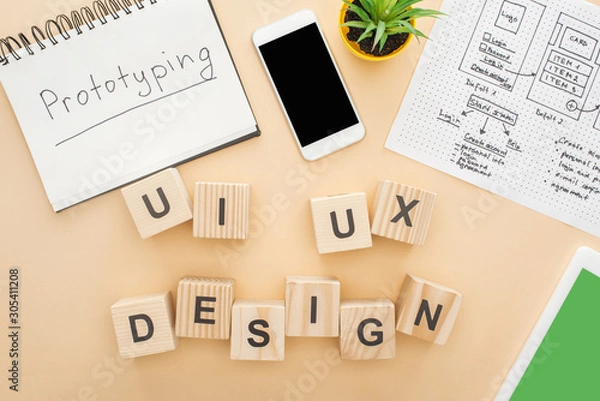 Fototapeta top view of gadgets near wooden blocks with ui and ux design lettering, website design template, notebook with prototyping lettering and green plant on beige background