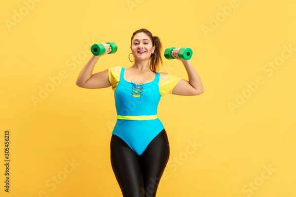 Fototapeta Young caucasian plus size female model's training on yellow background. Copyspace. Concept of sport, healthy lifestyle, body positive, fashion, style. Stylish woman emotional practicing with weights.