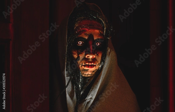 Fototapeta Creepy demonic witch in a hood with white eyes stands in front of the opened door