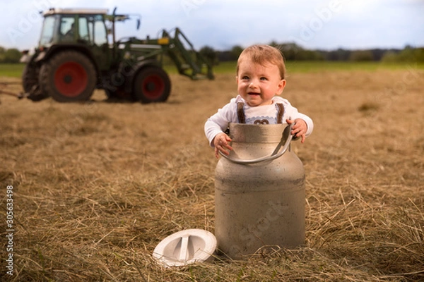 Obraz An adorably Bavarian baby boy standing in a milk churn laughing happily. In the background a tractor is turning  the hay the milk churn in standing on.