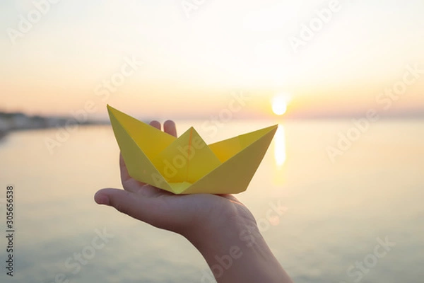 Obraz Closeup view of one hand of young kid holding paper yellow ship in hands isolated at sunny golden sunrise or sunset landscape. Horizontal color photography.