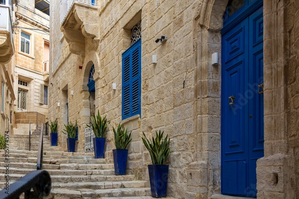 Fototapeta Narrow charming street in Birgu, Malta, with limestone medieval buildings with navy blue doors and window shutters and plants in blue pots along the walls.