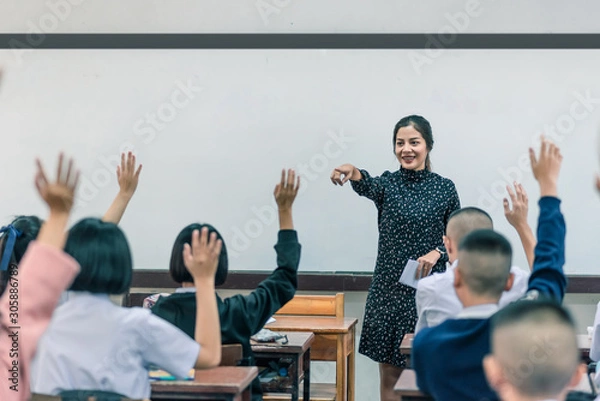 Fototapeta A smiling Asian female high school teacher teaches the white uniform students in the classroom by asking questions and then the students raise their hands for answers.
