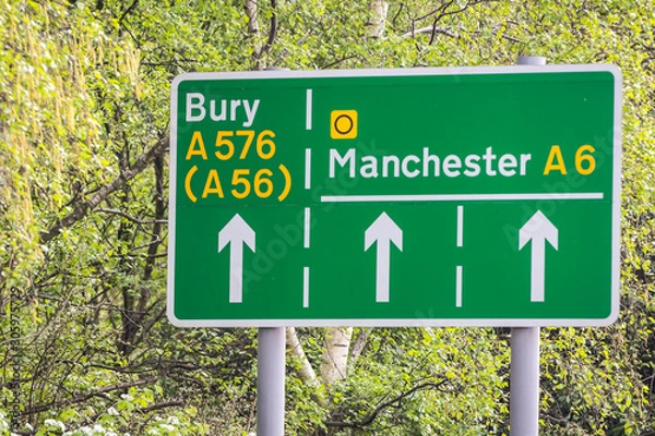 Obraz Green road signs on a leafy background showing directions to manchester and bury including the A6 A576 and A56.