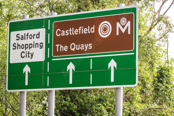 Obraz Green road signs on a leafy background showing directions to salford shopping city and and castlefield in greater manchester.