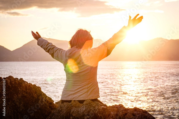 Fototapeta Woman sitting by ocean rejoicing with arms open wide to the sunset