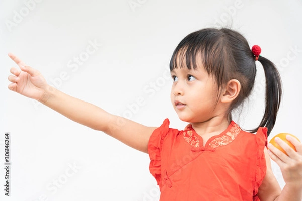 Obraz Adorable asian little girl with beautiful eyes in the red chinese dress is holding the orange and she is pointing on the right and smiling with fully happiness moment.