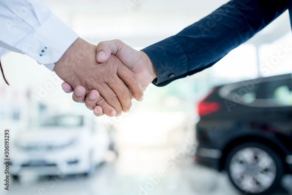 Obraz auto business, car sale, deal, gesture and people concept - close up of dealer giving key to new owner and shaking hands in auto show or salon