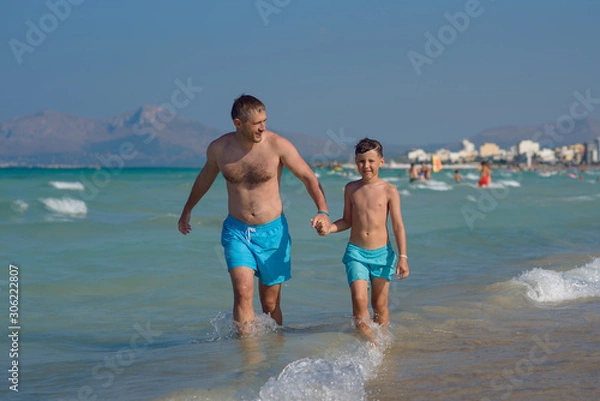 Fototapeta Dad and son are running in the water along the sea shore. They are holding hands and moving towards the camera.
