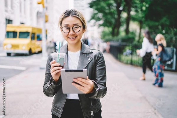Fototapeta Half length portrait of charming emotional asian female blogger enjoying recreation with digital tablet and tasty drink.Cute cheerful woman looking at camera and laughing while walking at street