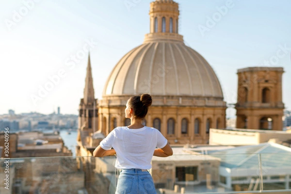 Fototapeta A brunette woman stands on a balcony at a height against the background of the city and the church with a dome. Focus on the woman. Back view