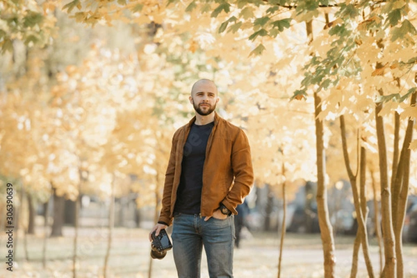 Obraz Bald brutal photographer with a beard in a suede leather jacket, blue shirt and jeans holds the camera and waits for a model in the park in the afternoon