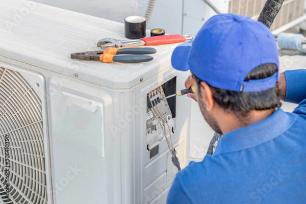 Fototapeta a professional electrician man is fixing a heavy duty unit of central air conditioning system by his tools on the roof top and wearing  uniform and white cap