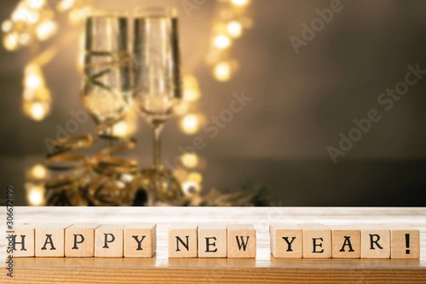 Fototapeta Happy New Year text made out of wooden blocks. Two glasses of champane with festive new year's eve decoration in the background. Copy space in the top right corner.