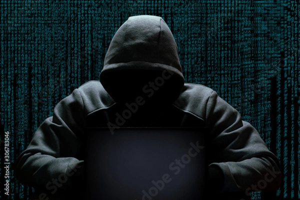 Fototapeta A cybercriminal in a hood without a face sits in the dark behind a laptop, against the backdrop of a binary code